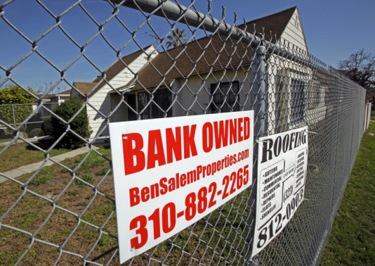 A vacant home surrounded by a chain link fence, carrying a "bank owned" sign.
